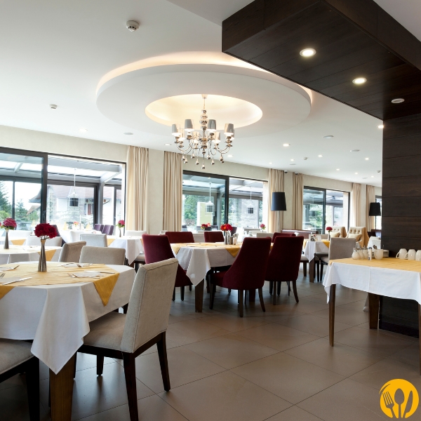 Jasna sala restauracyjna z dużymi oknami, białymi obrusami i okrągłym żyrandolem.