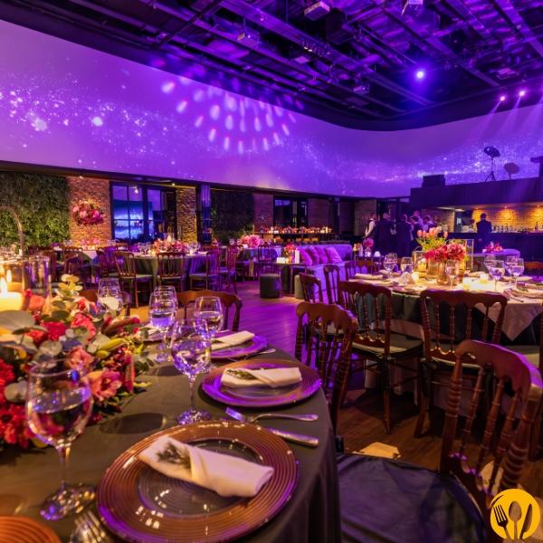 Sala bankietowa w fioletowym oświetleniu, nakryte stoły i dekoracje kwiatowe.